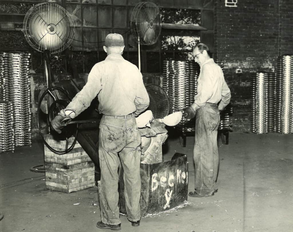 Historical photo of two molders pouring melted aluminum into a mold, with large fans blowing and neat stacks of cast aluminum parts in the background.