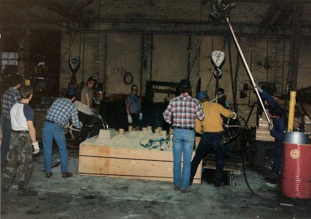 Historical photo inside Wisconsin Aluminum Foundry showing a crew of workers observing a molder pour melted aluminum into a very large air set mold while another worker holds a pulley system connected to the mold.