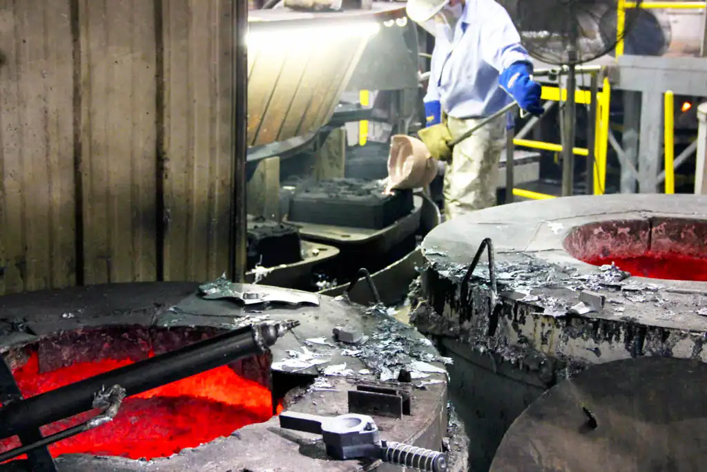 A working worker wearing protective attire ladles hot aluminum into mold.