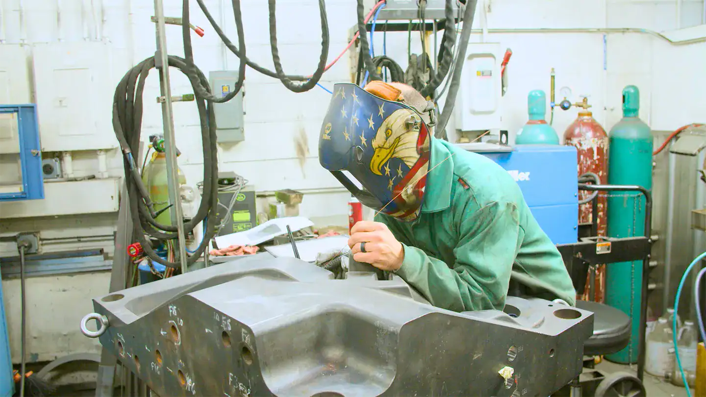 Worker in a manufacturing facility wearing a welding helmet leans over a. large aluminum part.