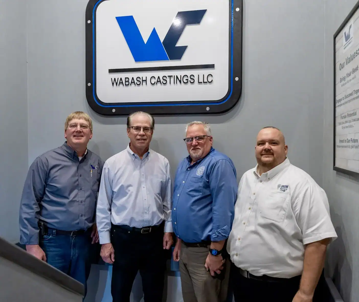 Indiana Senator Mike brown poses in front of the Wabash Castings wall logo aside three employees.