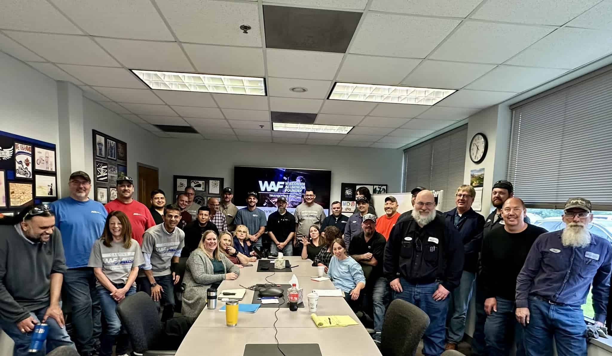 Conference room filled with smiling people, some wearing T-shirts with WAF and ATEK logos, with a presentation on screen in the background showing the Wisconsin Aluminum Foundry logo.