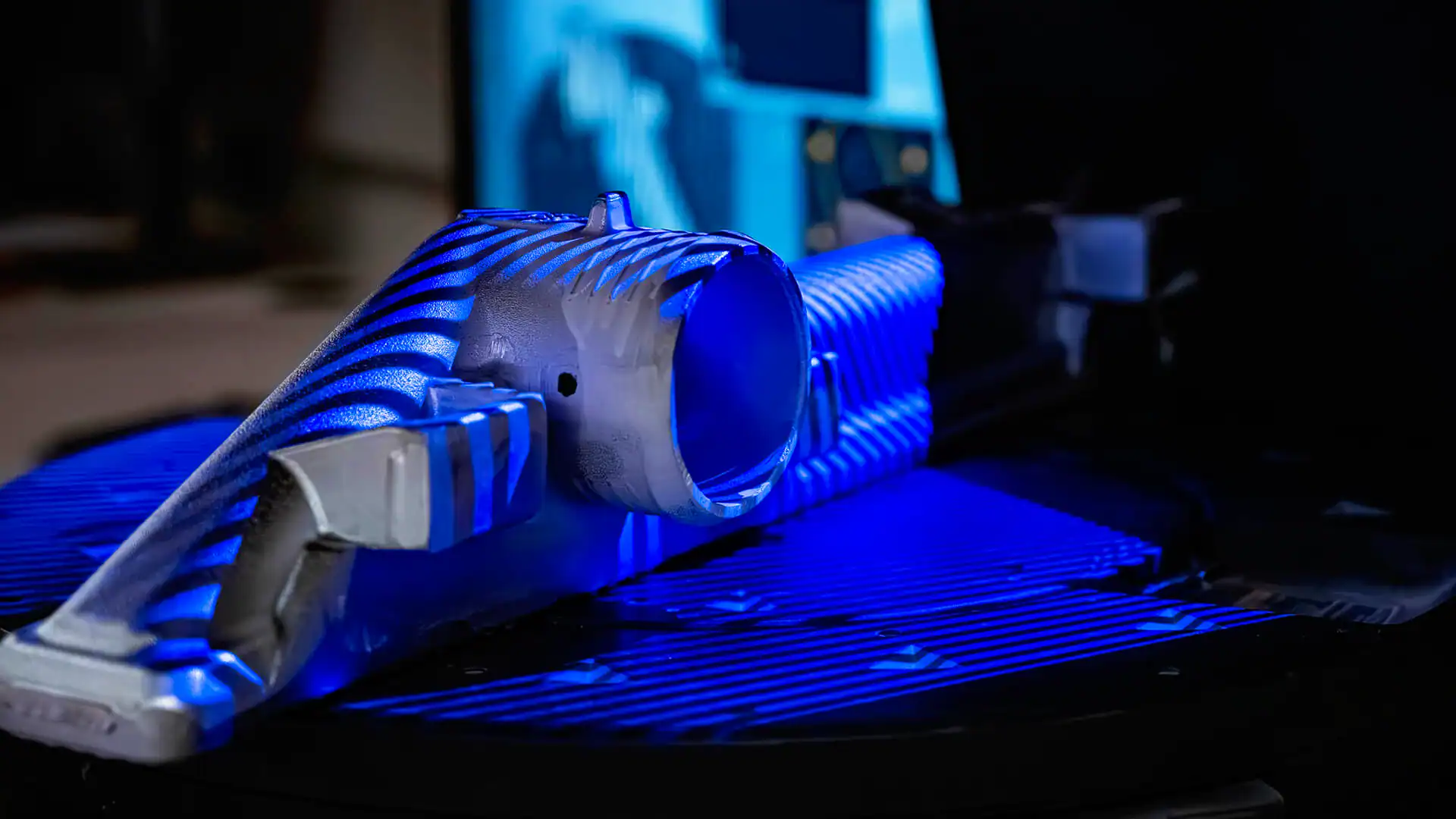 Closeup of small aluminum component under an X-ray inspection blue light scanner, with the glow of a screen in the background.
