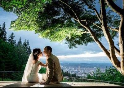 CineCrown Photography A bride and groom sitting on a bench overlooking a city.