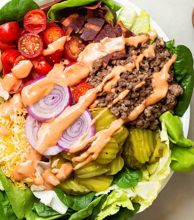 A big mac salad in a bowl with seasoned ground beef, grape tomatoes, red onions, pickles, turkey bacon and shredded cheese with special sauce