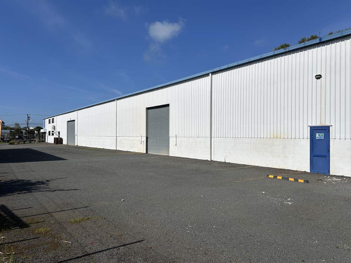 Large white industrial warehouse building with blue sky and parking area.
