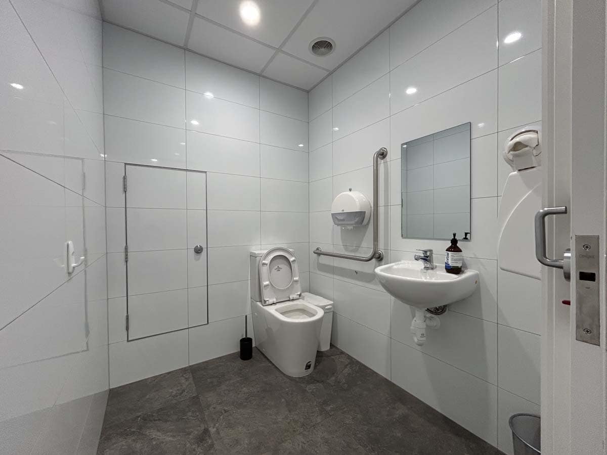 Clean and accessible public bathroom featuring white tiled walls, a toilet, and a sink with a mirror.