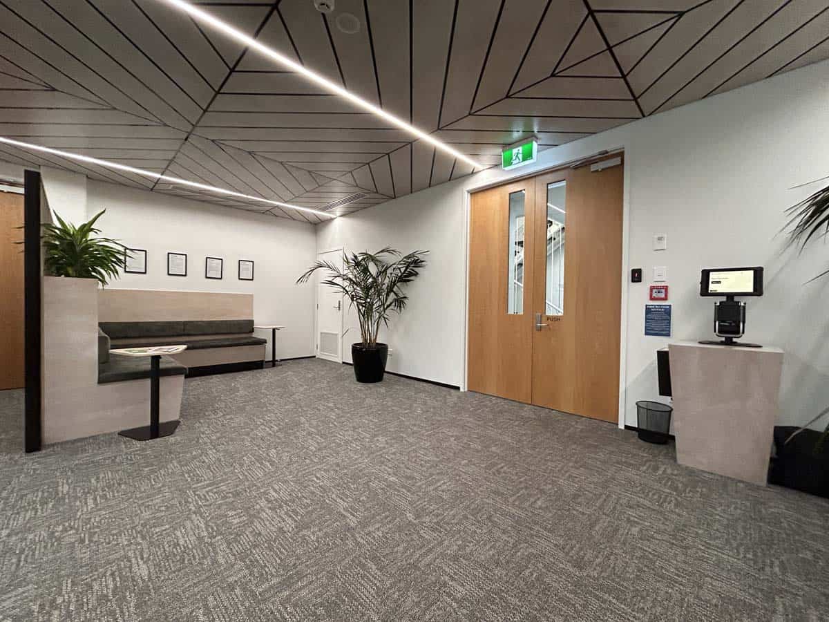 Modern office reception with seating, plants, and digital check-in kiosk.