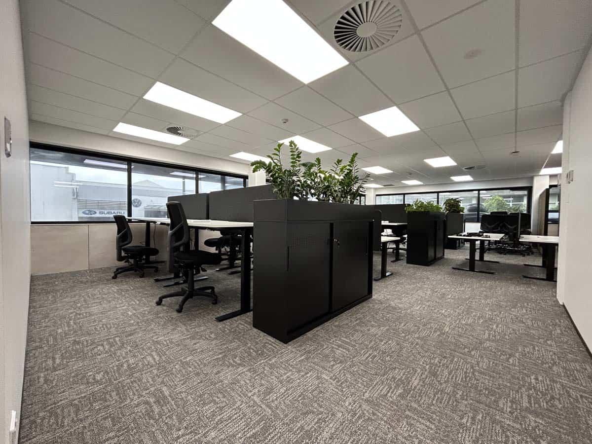 Modern office workspace with desks, chairs, and plants in a bright, open-plan environment.