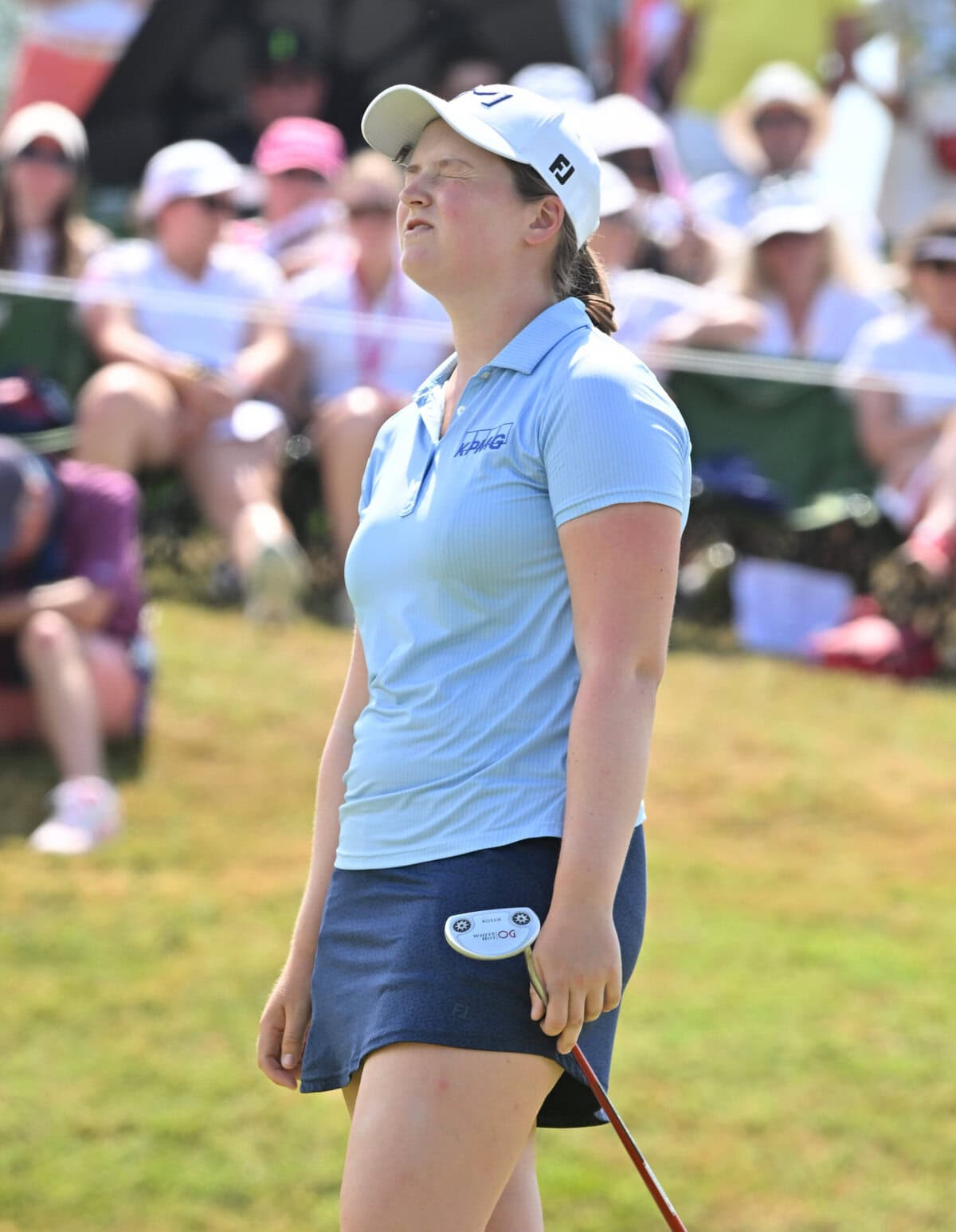 Lottie Woad stands on the green with her eyes closed and head tilted upward, holding her putter as she reacts in visible frustration during the Evian Championship, with spectators watching behind her.