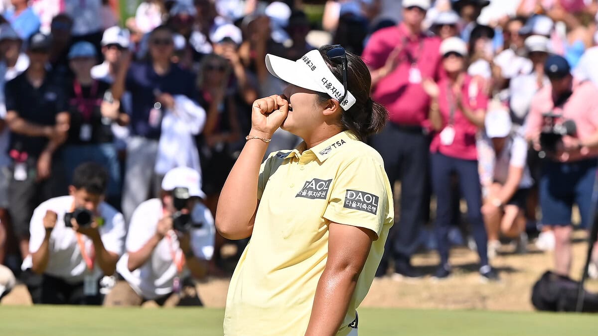 Haeran Ryu stands on the green in a yellow shirt and white visor, holding her club and reacting emotionally as a large crowd of spectators and photographers applaud her victory at the Evian Championship.