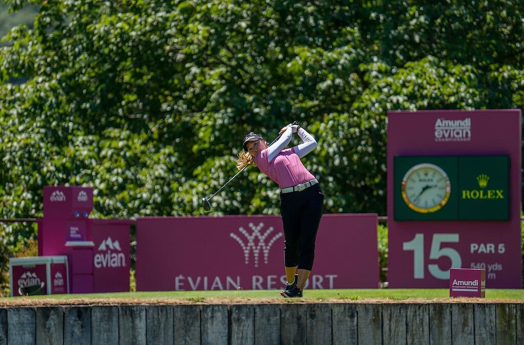 Brooke Henderson tees off on the 15th hole at the Amundi Evian Championship, wearing a pink shirt and cap, with tournament signage and a Rolex clock behind her as she drives the ball from the tee box.