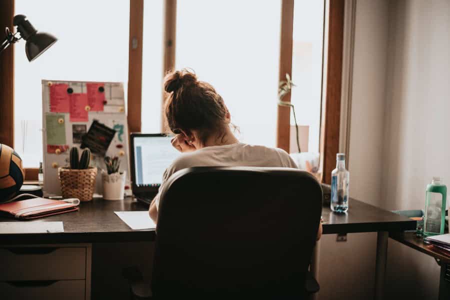 Young woman working at a cluttered home desk, focused on her laptop in a small workspace.