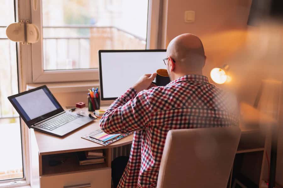 Man in a checked shirt working from a home office desk with dual screens and a laptop.