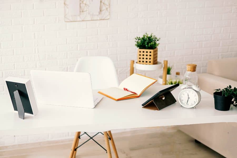 Bright minimalist white desk with laptop, notebook, clock, plants and decor arranged neatly.