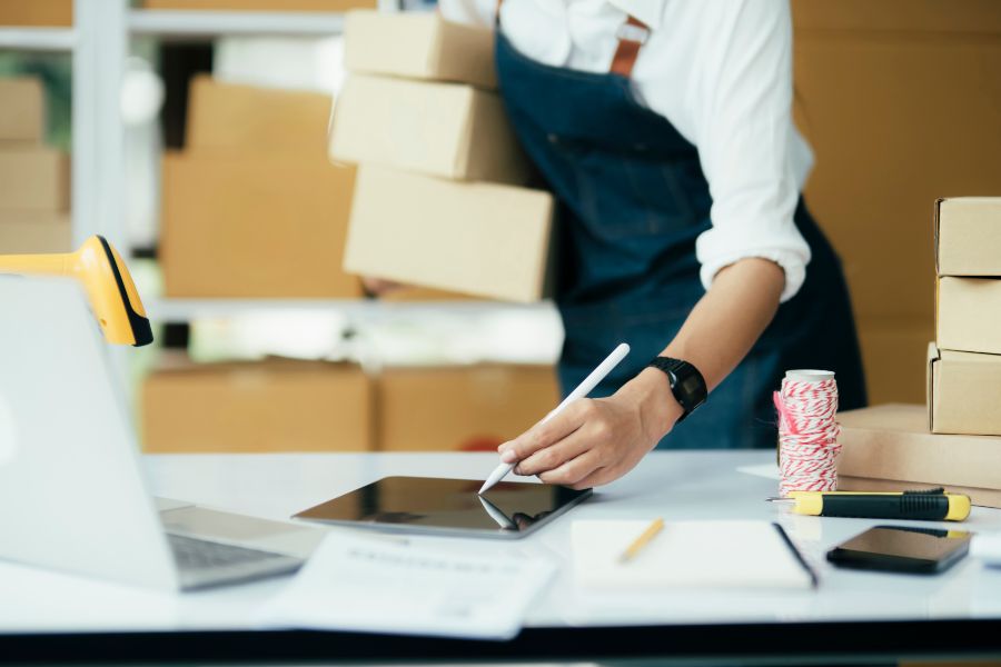 A person working with a tablet and shipping boxes in a small business inventory setting.