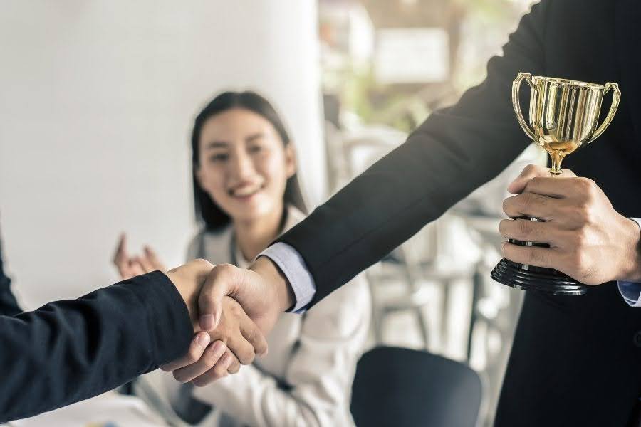 Two professionals shaking hands over a trophy, representing pay and entitlements when hiring your first employee