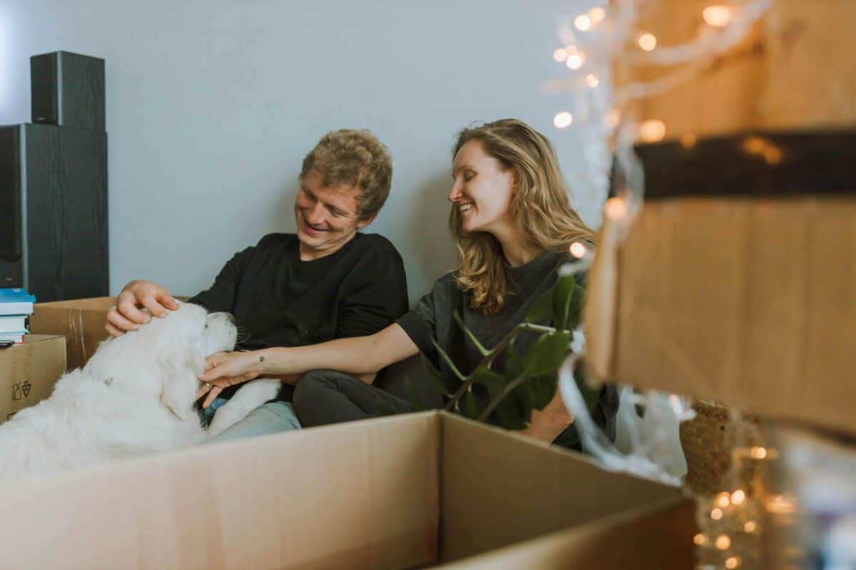 A smiling couple sits on the floor of their new home surrounded by moving boxes, while their dog nudges them for attention. This image represents the comfort and support offered through corporate pet relocation.