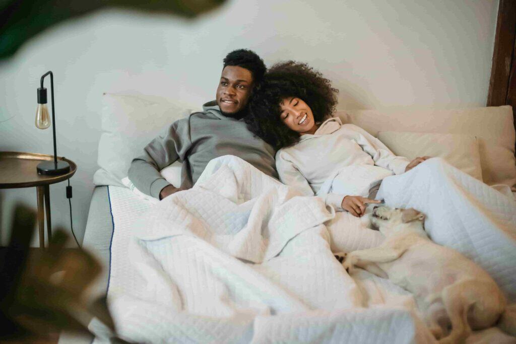 Happy ethnic couple lying in bed with dog during lazy weekend at home.
