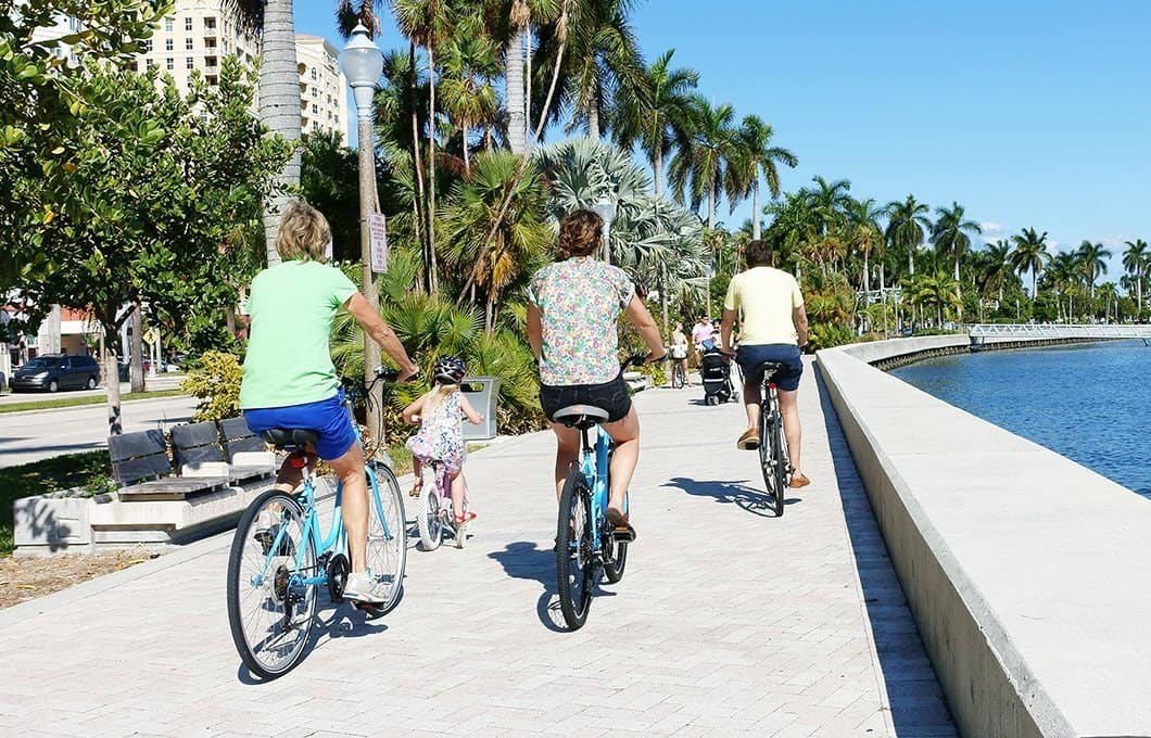 People cycling along a waterfront promenade with palm trees and city buildings in the background.