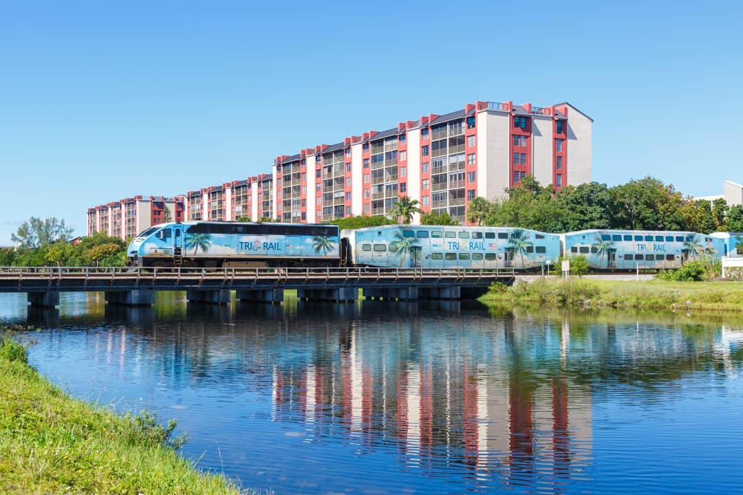 Modern train passing by a waterway with a large apartment building in the background.