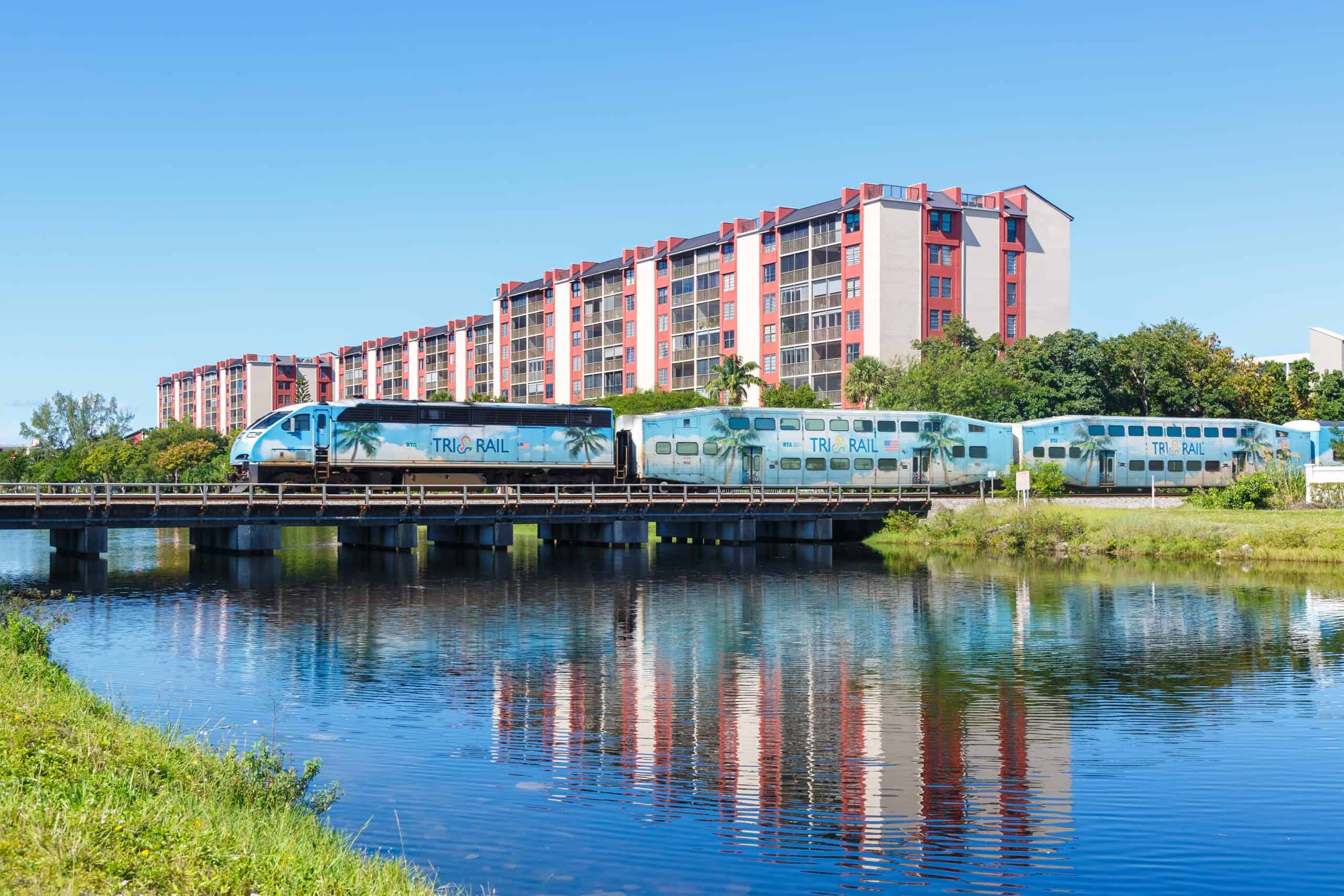 Modern train passing by a waterway with a large apartment building in the background.