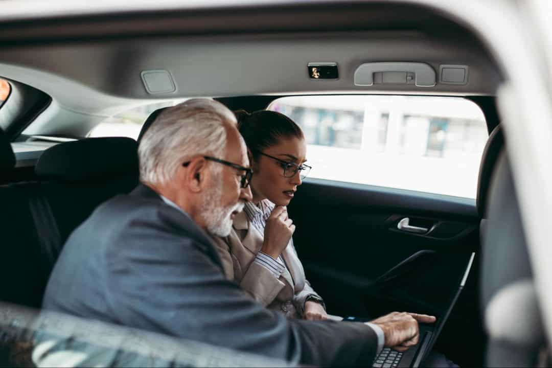 An older gentleman describes what he sees on a laptop screen to a younger executive while carpooling in a vehicle.