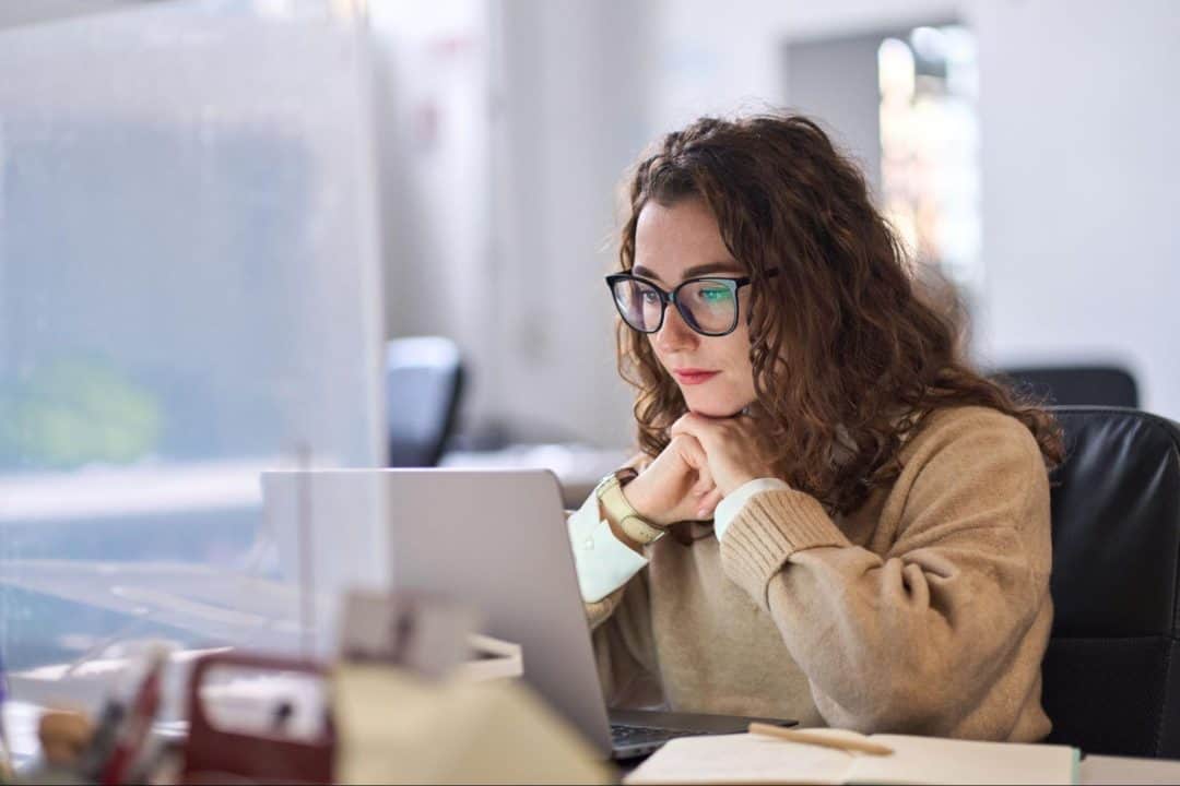 A young professional woman in glasses, contemplating choices while sitting at her desk in front of her laptop.