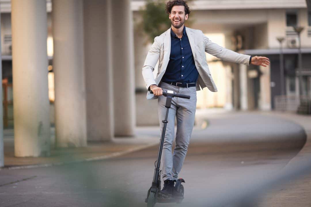 A businessman with a white jacket and grey slacks rides an electric scooter to his destination.