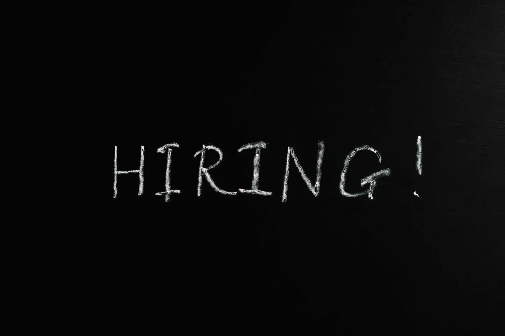 The word 'HIRING!' is written in large, rough white chalk letters on a blackboard, emphasizing job recruitment and employment opportunities. This simple yet bold message reflects the evolving landscape of job hiring in the digital age, where Artificial Intelligence (AI) is changing the job market by automating recruitment processes, optimizing candidate selection, and transforming how companies fill open positions. The stark contrast between the white chalk and dark background highlights the urgency and demand for skilled professionals in an AI-driven workforce.