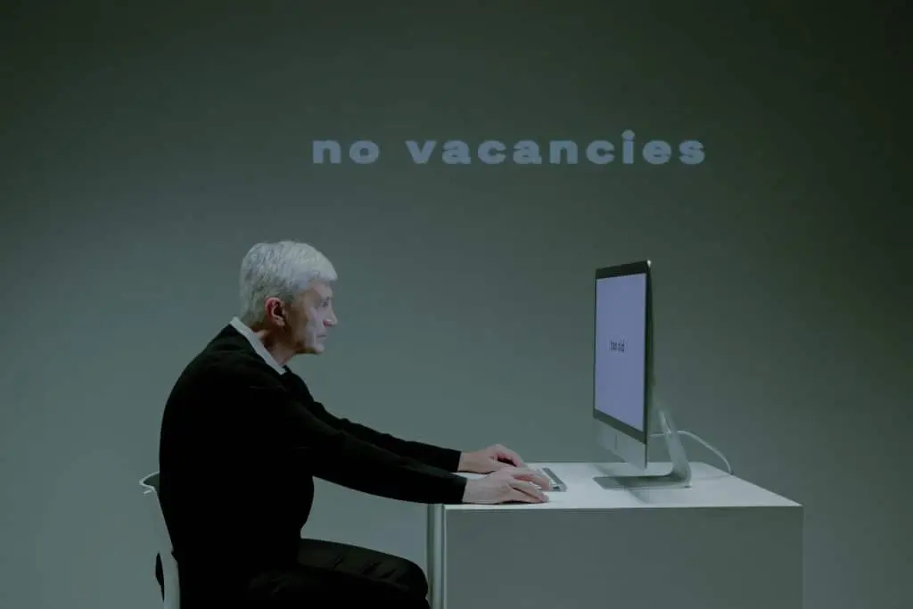 An elderly man with gray hair and a black sweater sits at a modern white desk, typing on a computer. The screen displays the words 'too old,' while a large projected text behind him reads 'no vacancies.' The dim lighting and minimalist setting emphasize the growing concern about how Artificial Intelligence (AI) is changing the job market, particularly in terms of hiring biases and automated decision-making. This thought-provoking image highlights AI’s role in employment discrimination and challenges older job seekers face in an evolving workforce.