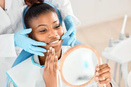 woman sitting in dental chair, looking at teeth in mirror, dentists hands around her face
