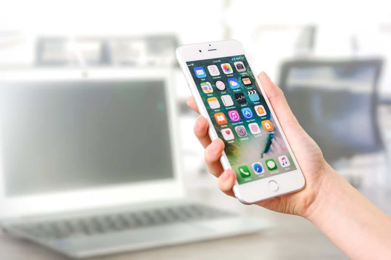 hero-about Close-up view of a smartphone displaying apps, held by a hand, with a blurred laptop in the background.