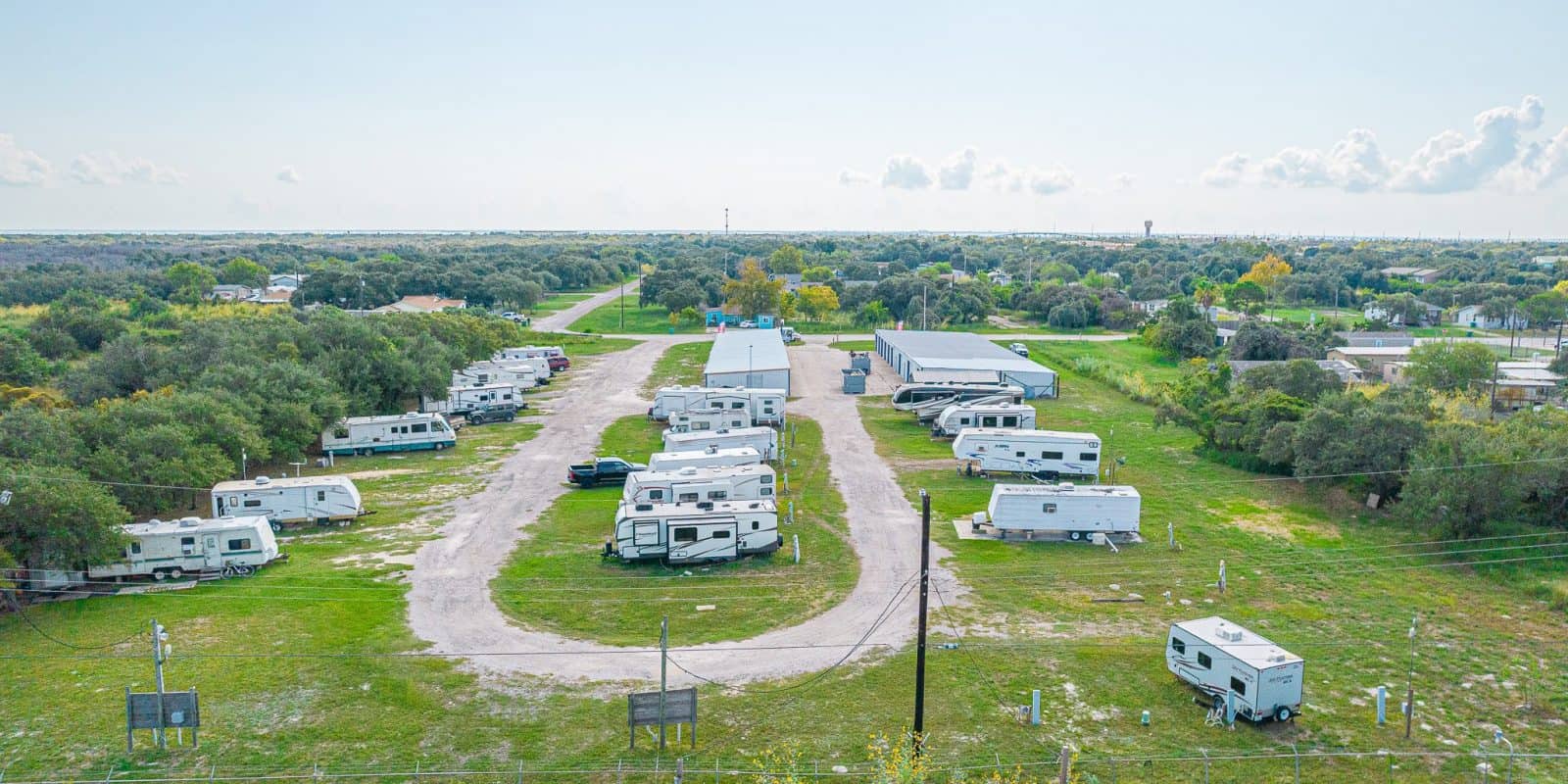 Aransas Pass RV Park and Storage Aerial view of Aransas Pass RV and Storage park with spacious RV sites, white campers, curved gravel road, storage units, green trees.