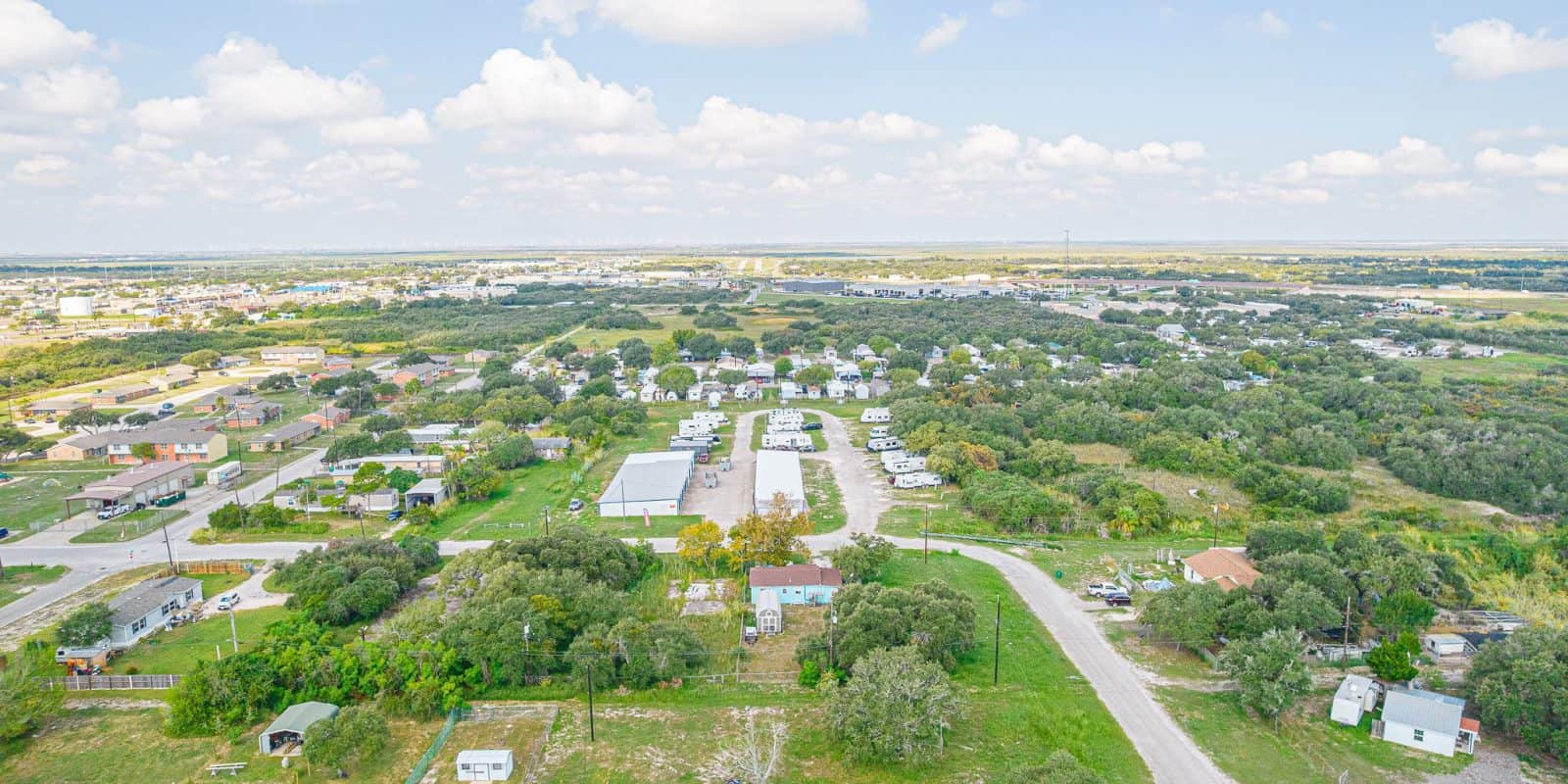 Aransas Pass RV Park and Storage Aerial view of Aransas Pass RV and Storage park with white RVs, houses, trees, curved roads, and open fields under cloudy sky.