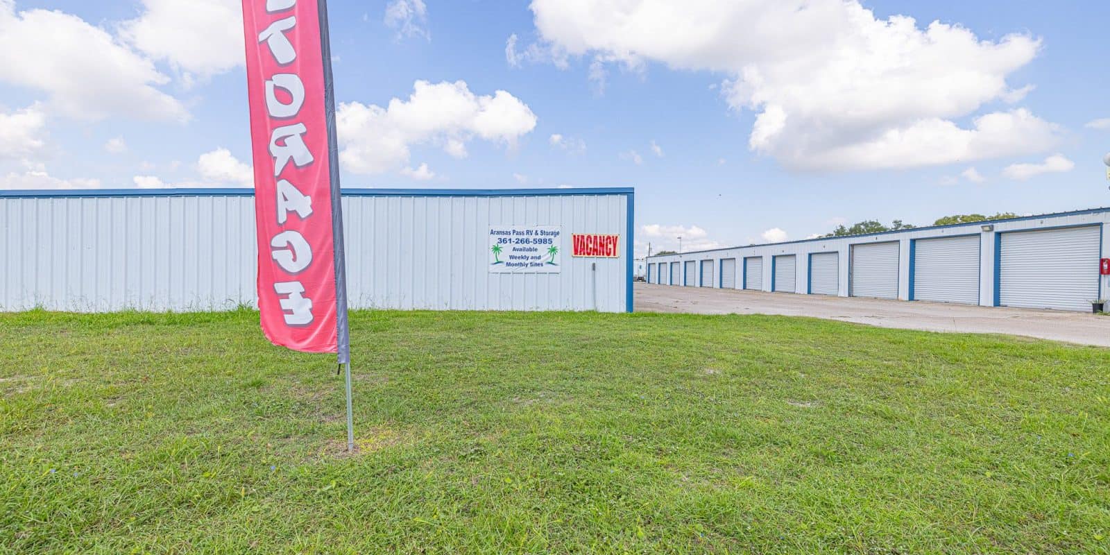 Aransas Pass RV Park and Storage Aransas Pass RV and Storage blue units with grassy front, red STORAGE flag, business banners, drive-through & back-in spaces.