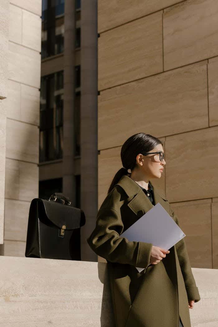 about-us Businesswoman in a coat holding documents outside an office building with a briefcase nearby.