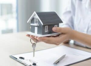 Une personne portant une chemise blanche tient une petite maison modèle et une clé – une image qui capture le processus immobilier au Québec, impliquant peut-être un courtisan préparant des documents et une soumission pour l'achat d'une maison sur une table en bois clair.