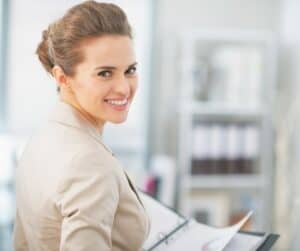 A woman with brown hair in an updo, wearing a beige blazer, smiles at the camera while holding an open binder—perhaps discussing assurance options or prix details—in a bright office with shelves of books and folders in the blurred background.