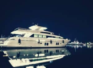A large white luxury yacht is docked at a marina at night, illuminated by soft lights. Its reflection shimmers in the calm water. Like a comparateur for prix, other boats and distant lights glimmer under the dark, starry sky.