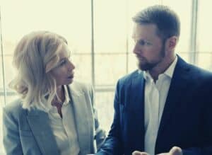 A woman and a man in business attire stand indoors by large sunlit windows, engaged in a serious conversation. The contemplative mood suggests they might be discussing a comparateur de prix for a project in Quebec.