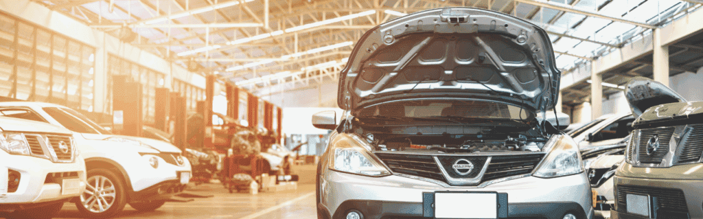 Une Nissan argentée, capot ouvert, est garée dans un atelier spacieux et lumineux. Pendant que les mécaniciens travaillent, un comparateur permet aux clients de trouver le meilleur prix pour leurs réparations dans un intérieur industriel et ensoleillé.