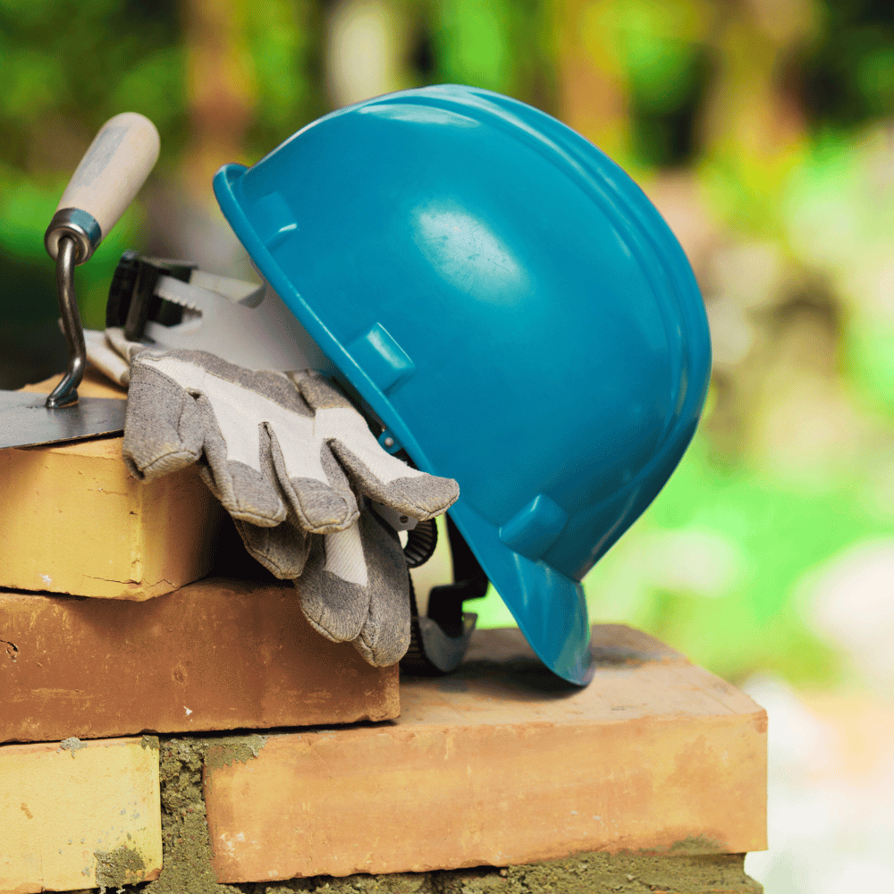 Quote Masonry Business Insurance A blue hard hat, grey work gloves and a trowel rest on a pile of yellow and red bricks. The fuzzy green foliage suggests an outdoor construction site: a perfect image for a masonry company or a quick quote.