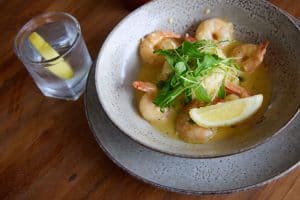 Succulent prawn dish with lemon and fresh greens in a rustic ceramic bowl, served with sparkling water and lemon wedge.