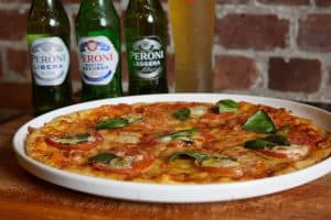 Bella Vita Italian pizza with fresh tomatoes, mozzarella, basil, served with Peroni beer bottles in background.