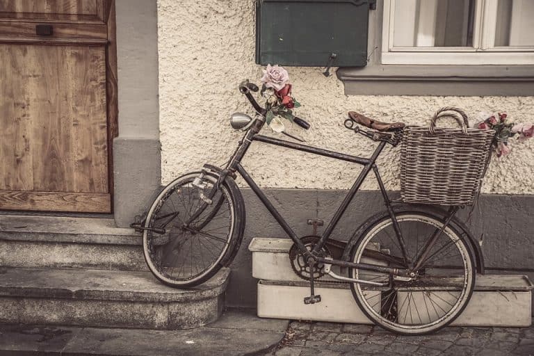 Bici che amore, bicicletta vintage con cestino di vimini decorato con fiori, posizionata davanti a una casa con parete bianca e porta in legno. Perfetta per una passeggiata romantica e nostalgica.