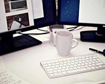 Computer workspace with dual monitors displaying documents and images, white keyboard, mug, notepad, pen, and other office essentials on a clean white desk.