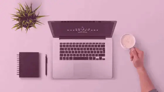 Laptop with open screen, notebook, pen, and a hand holding a coffee cup on a pink surface.