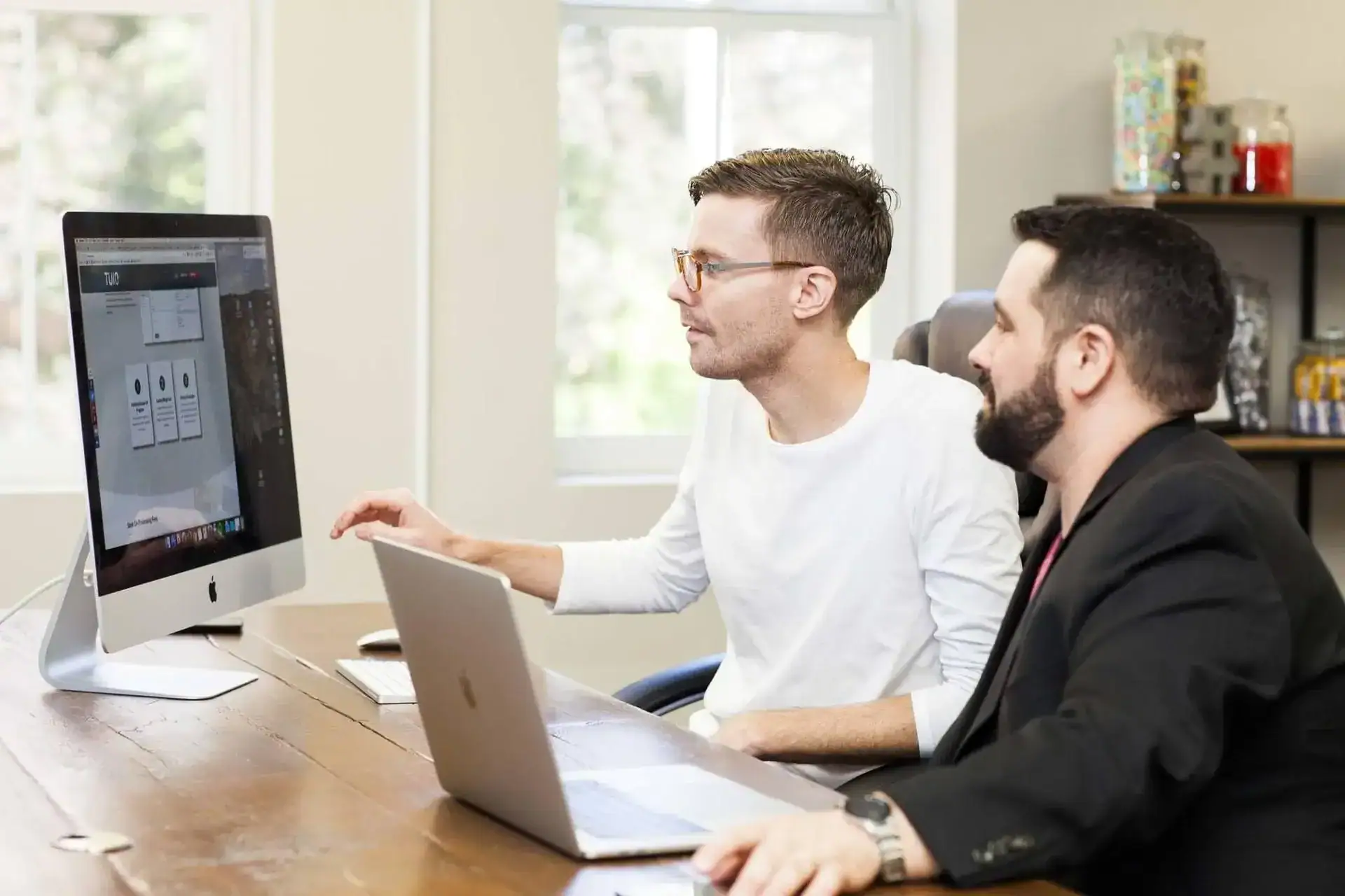 An image of two professionals collaborating on a project using a desktop computer and a laptop in a.