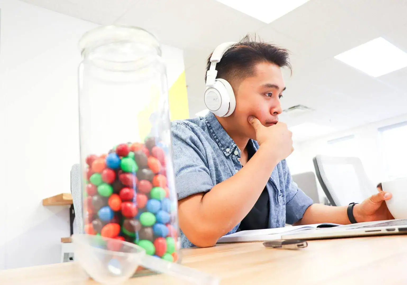 Young man wearing headphones, sitting at a desk with a jar of colorful gumballs.